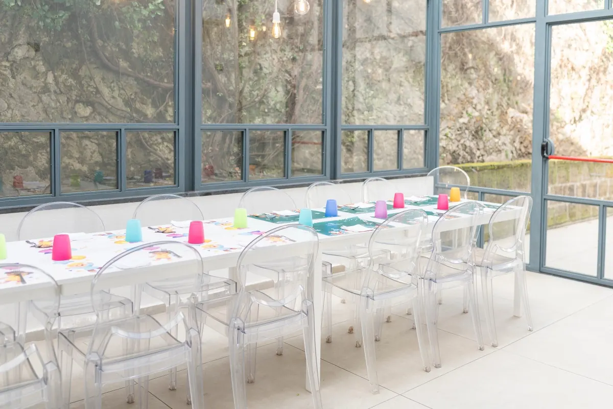 Baby’s Room vetrata con un lungo tavolo apparecchiato per i bambini, sedie trasparenti e bicchieri colorati