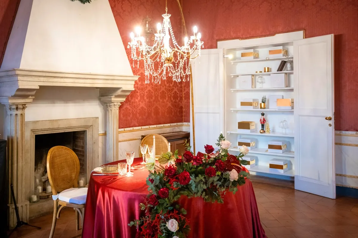 Salotto damascato rosso con camino, lampadario di cristallo e tavola rotonda in raso rosso con composizione di rose