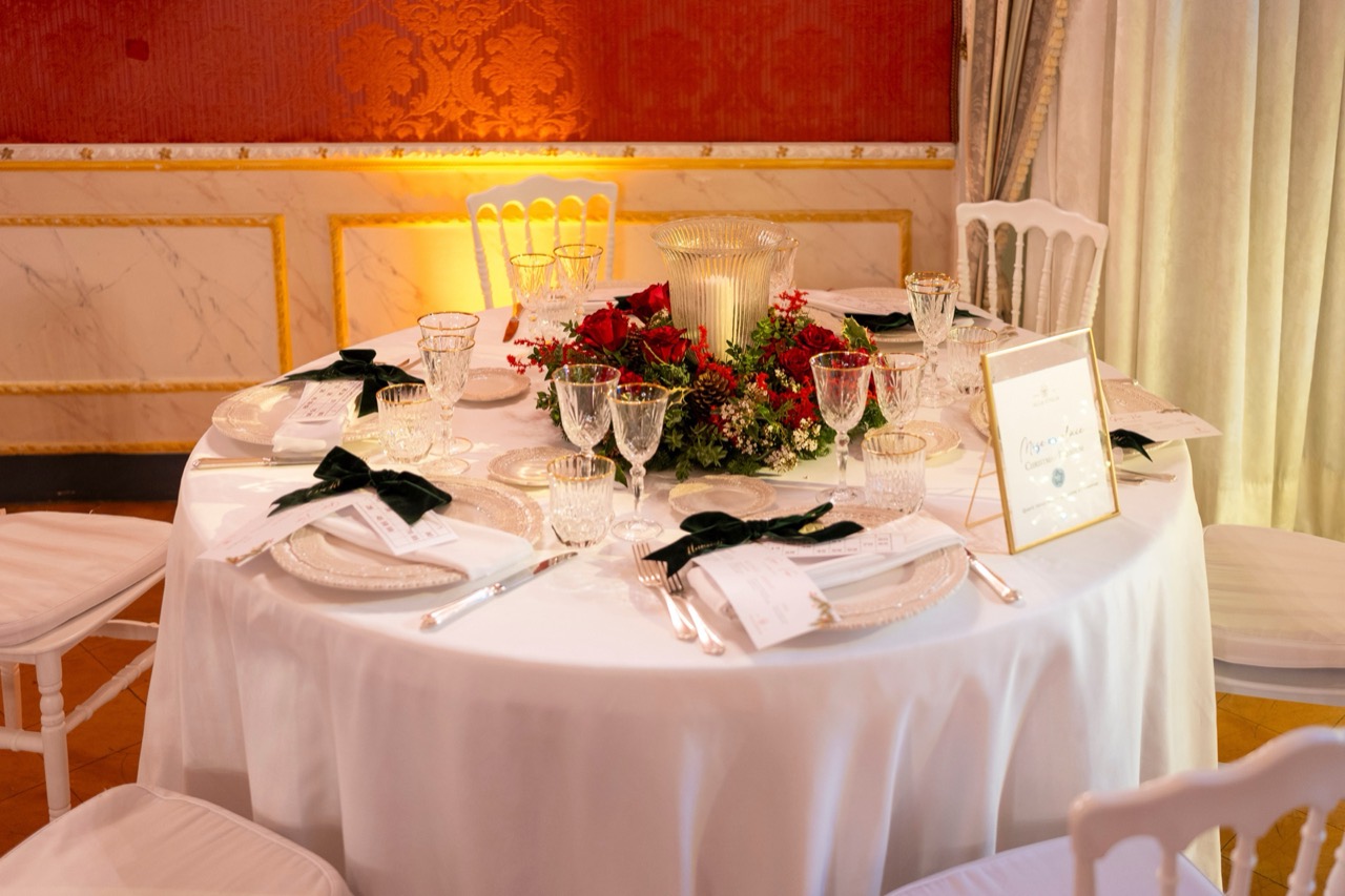 Tavolo rotondo apparecchiato per Natale con centrotavola di rose rosse e bacche, calici molati e fiocchi di velluto verde, nel salotto damascato rosso