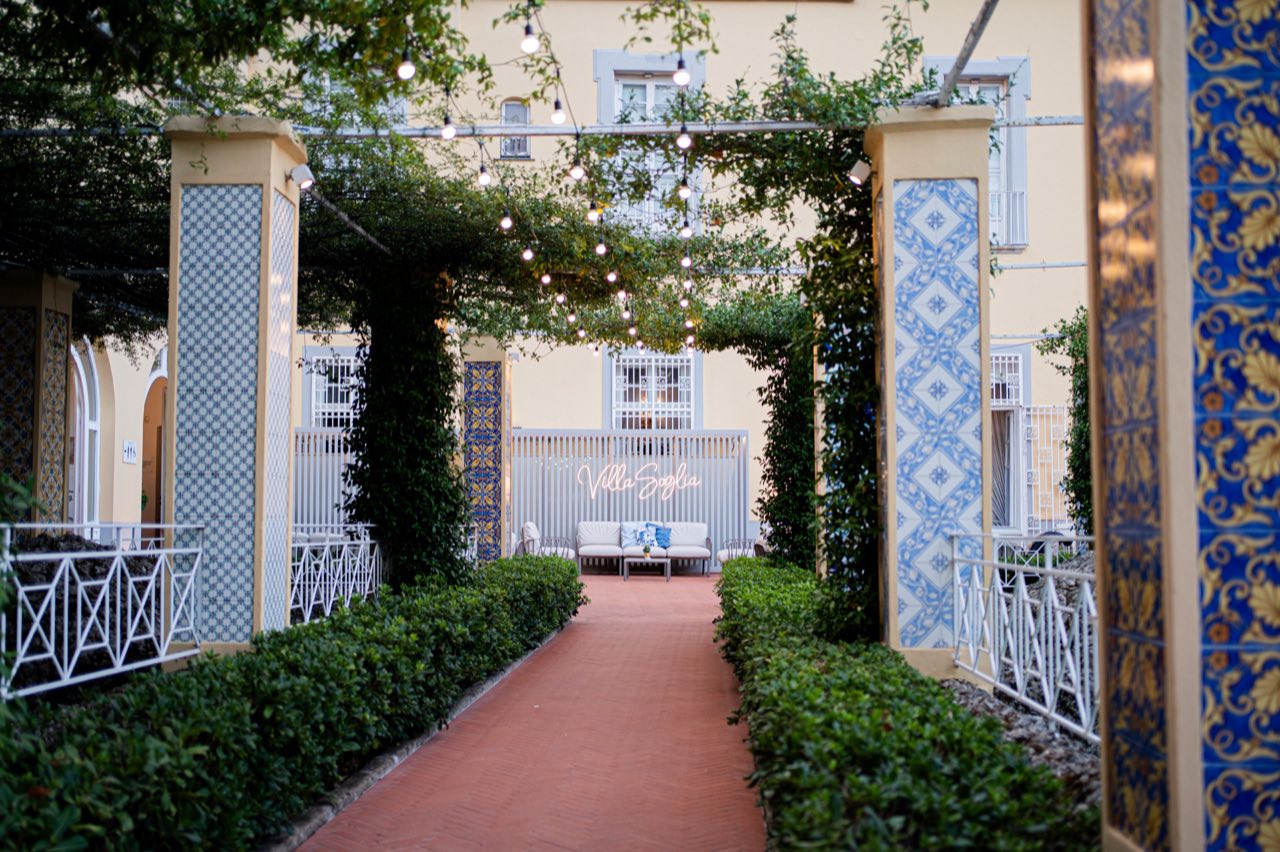 Vialetto sotto il pergolato fra pilastri di maioliche bianche e blu, con il salotto all’aperto e l’insegna Villa Soglia in fondo
