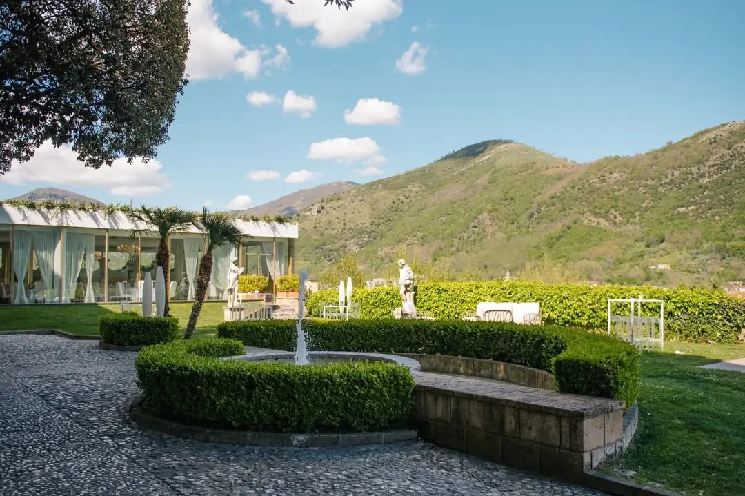 Prato d’ingresso con aiuole di bosso attorno alla fontana, la sala vetrata sullo sfondo e le colline della valle