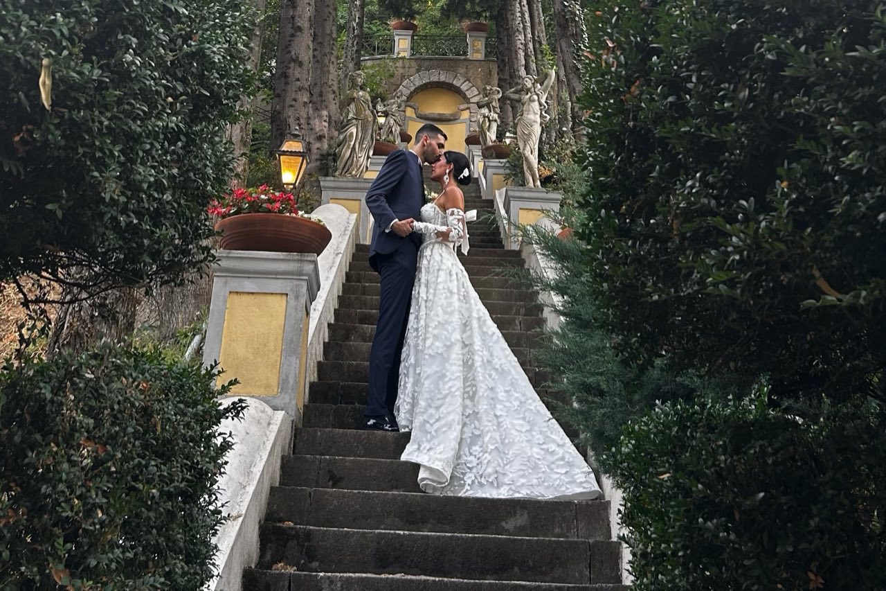 Sposi che si baciano sulla scalinata del parco tra le siepi, con statue, lanterna e fontana in cima