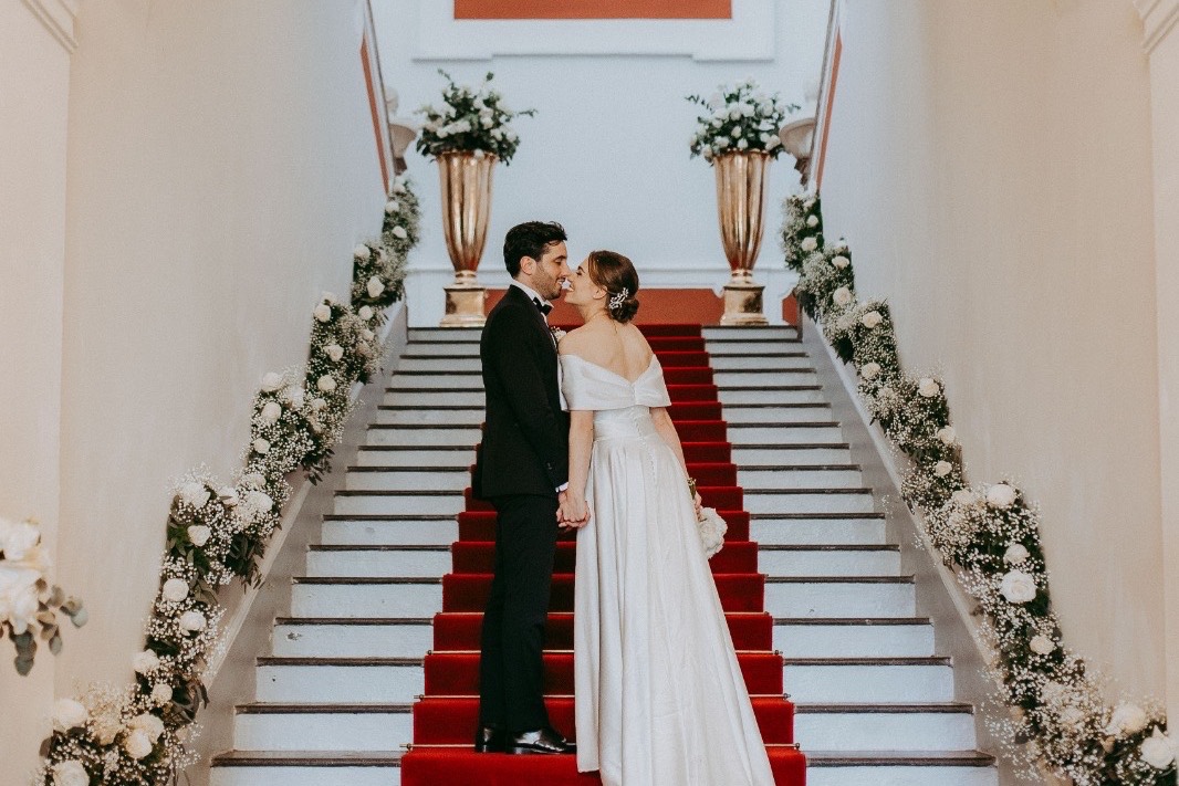 Sposi mano nella mano sulla passatoia rossa dello scalone d'onore, fra ghirlande di rose bianche e vasi dorati