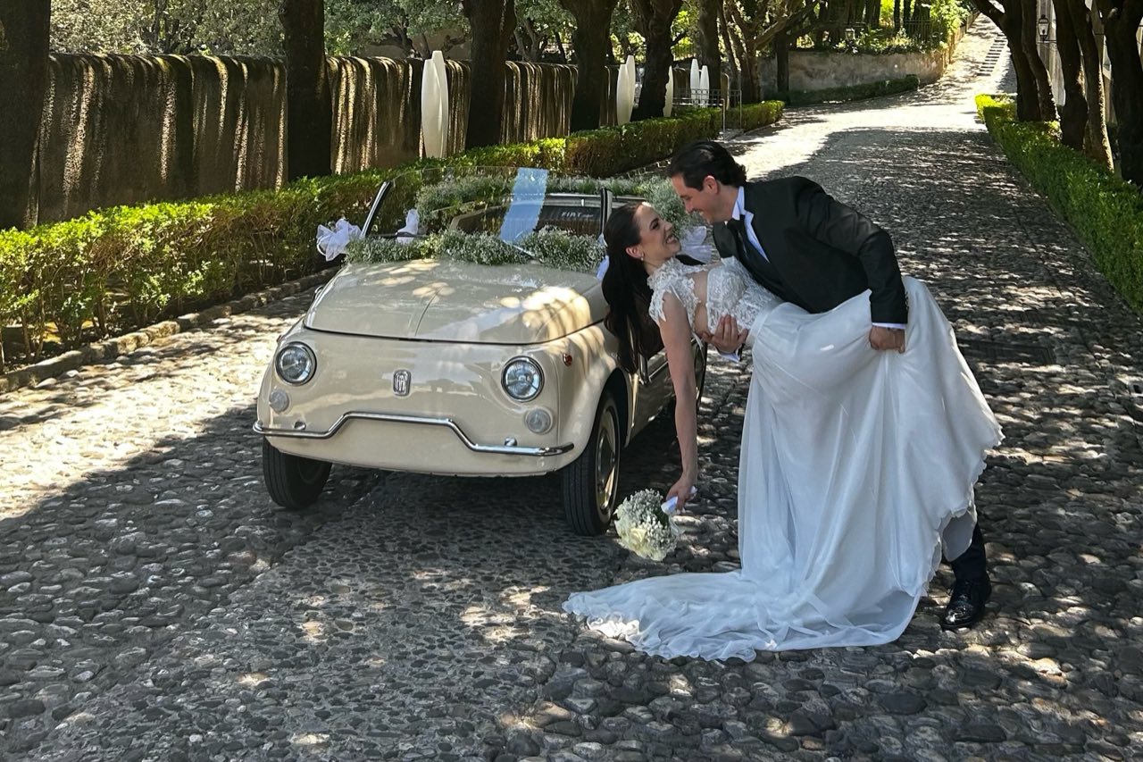 Sposo che fa il casqué alla sposa accanto a una Fiat 500 d'epoca decorata, nel viale acciottolato fra i lecci