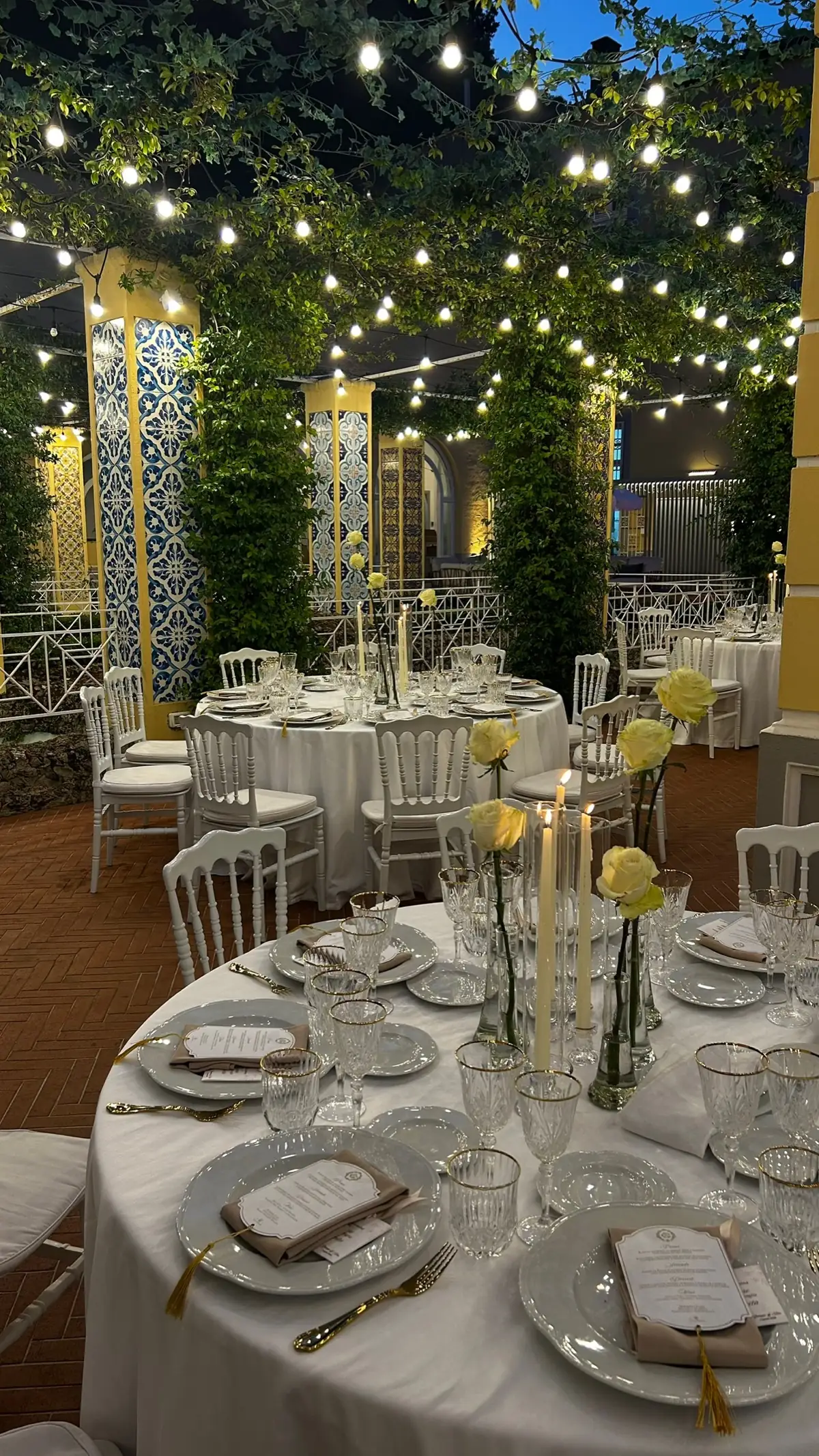 Tavoli rotondi con fiori gialli sotto le luci sospese del colonnato, fra i pilastri di maioliche