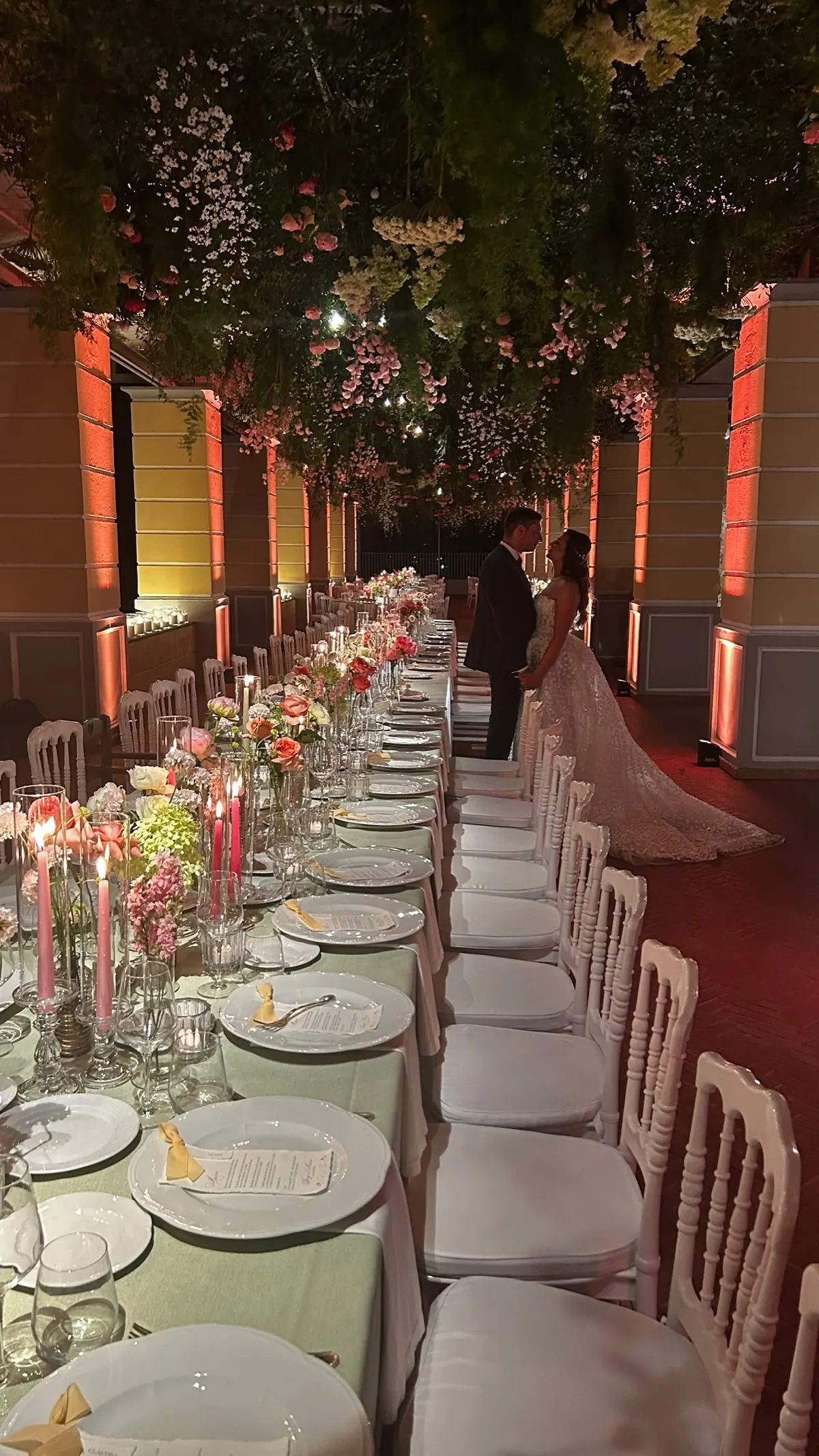 Tavolata imperiale con candele rosa sotto un soffitto di fiori, con gli sposi in fondo al colonnato