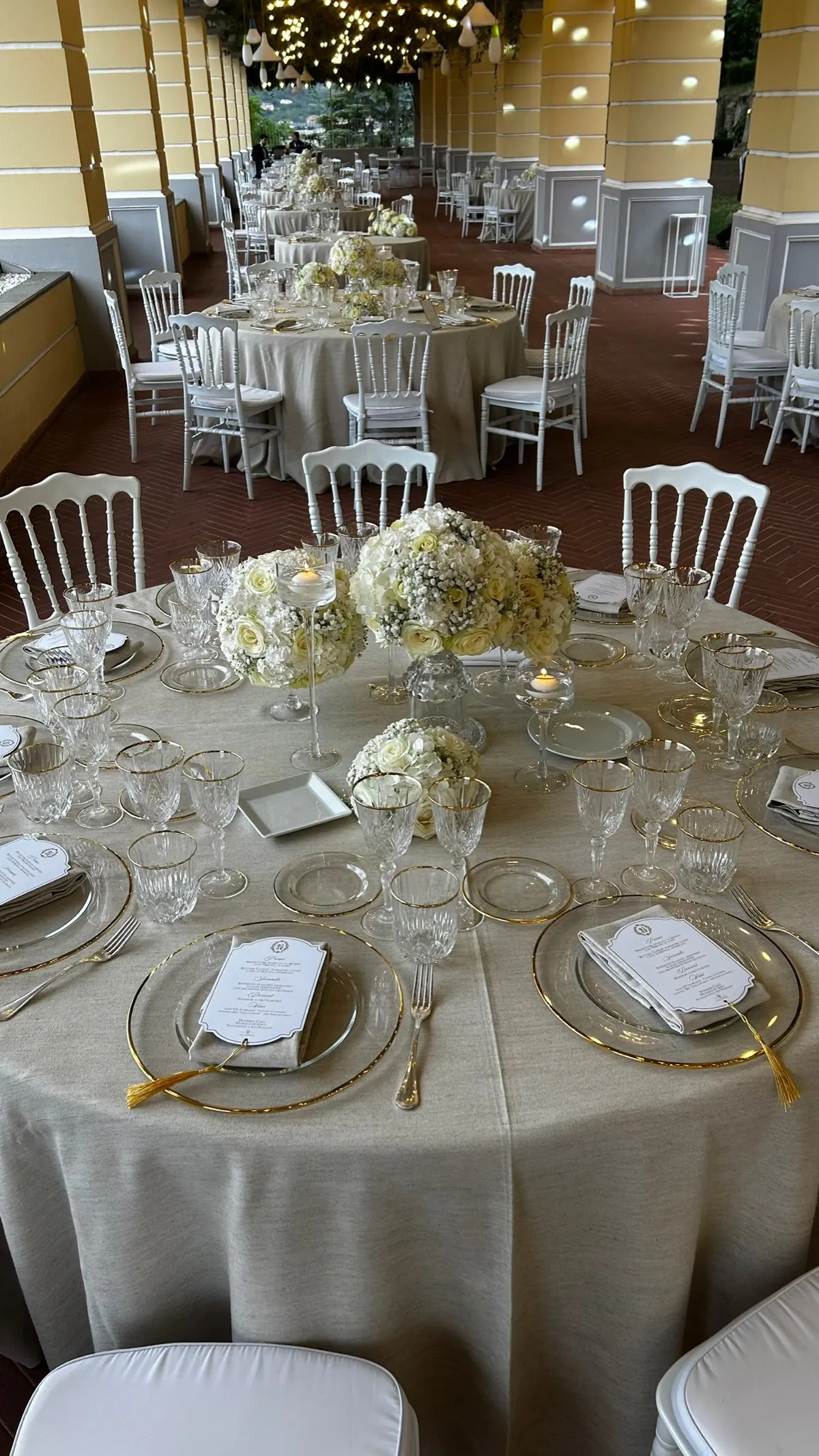 Tavoli rotondi sotto il colonnato della Sala Garden Glamour, con sottopiatti dorati, menu e centrotavola di fiori bianchi