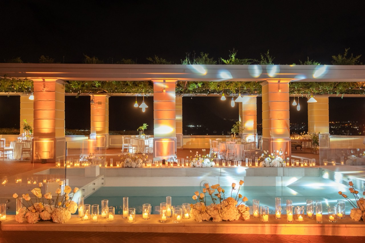 Piscina bordata di candele e rose bianche con il colonnato illuminato e la vista sulla valle