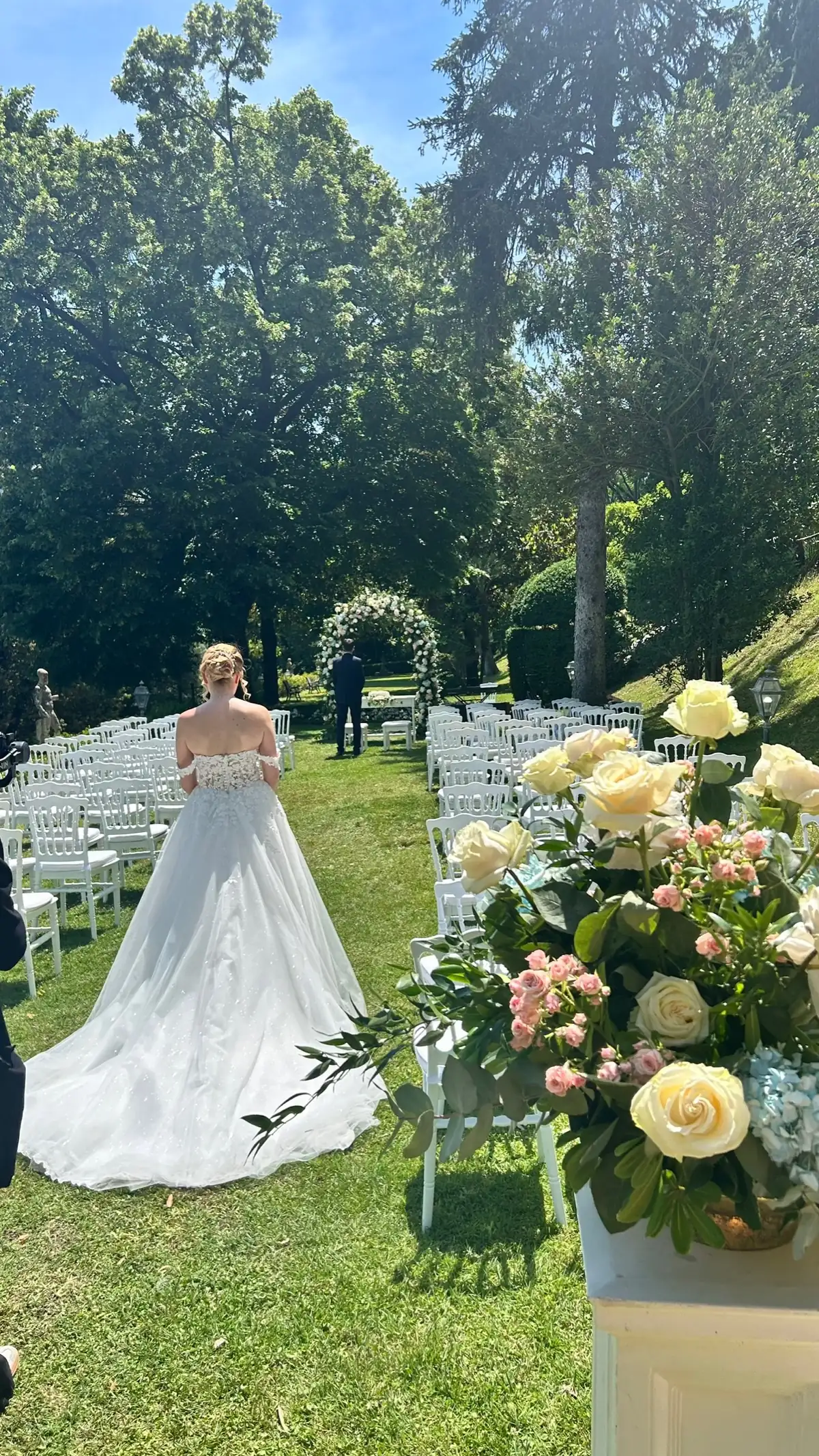 Ingresso della sposa nel rito civile in giardino, fra le sedute bianche e le composizioni di rose