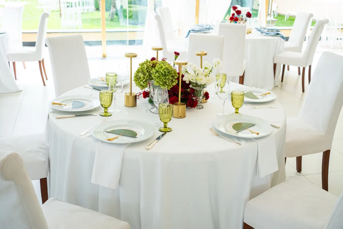 Tavolo rotondo con calici verdi, candele dorate, ortensie e rose rosse nella Sala Ricevimenti, con il prato oltre le vetrate