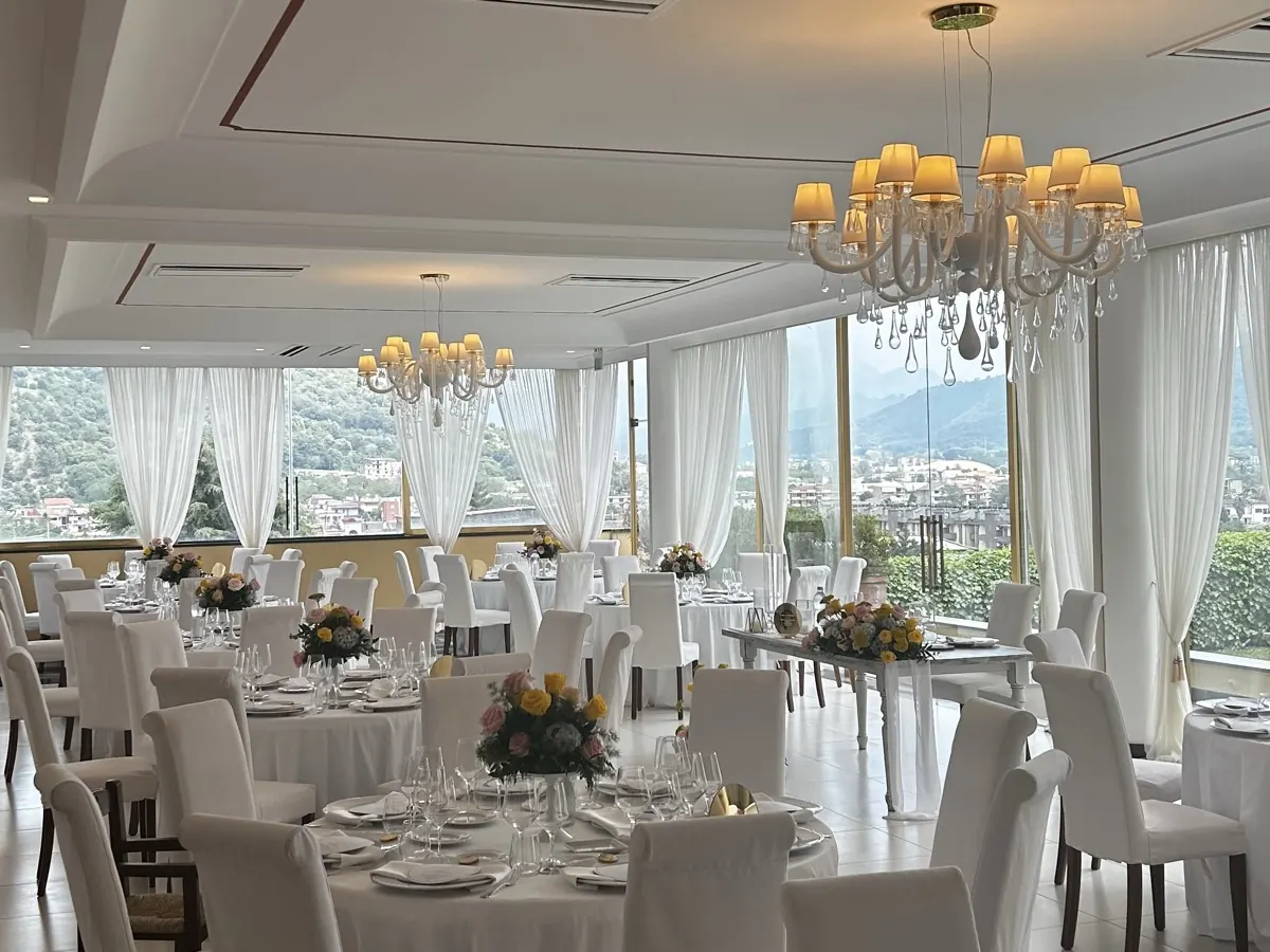 Sala Ricevimenti apparecchiata con tavoli rotondi e sedie vestite di bianco, lampadari di cristallo e vetrate sui monti