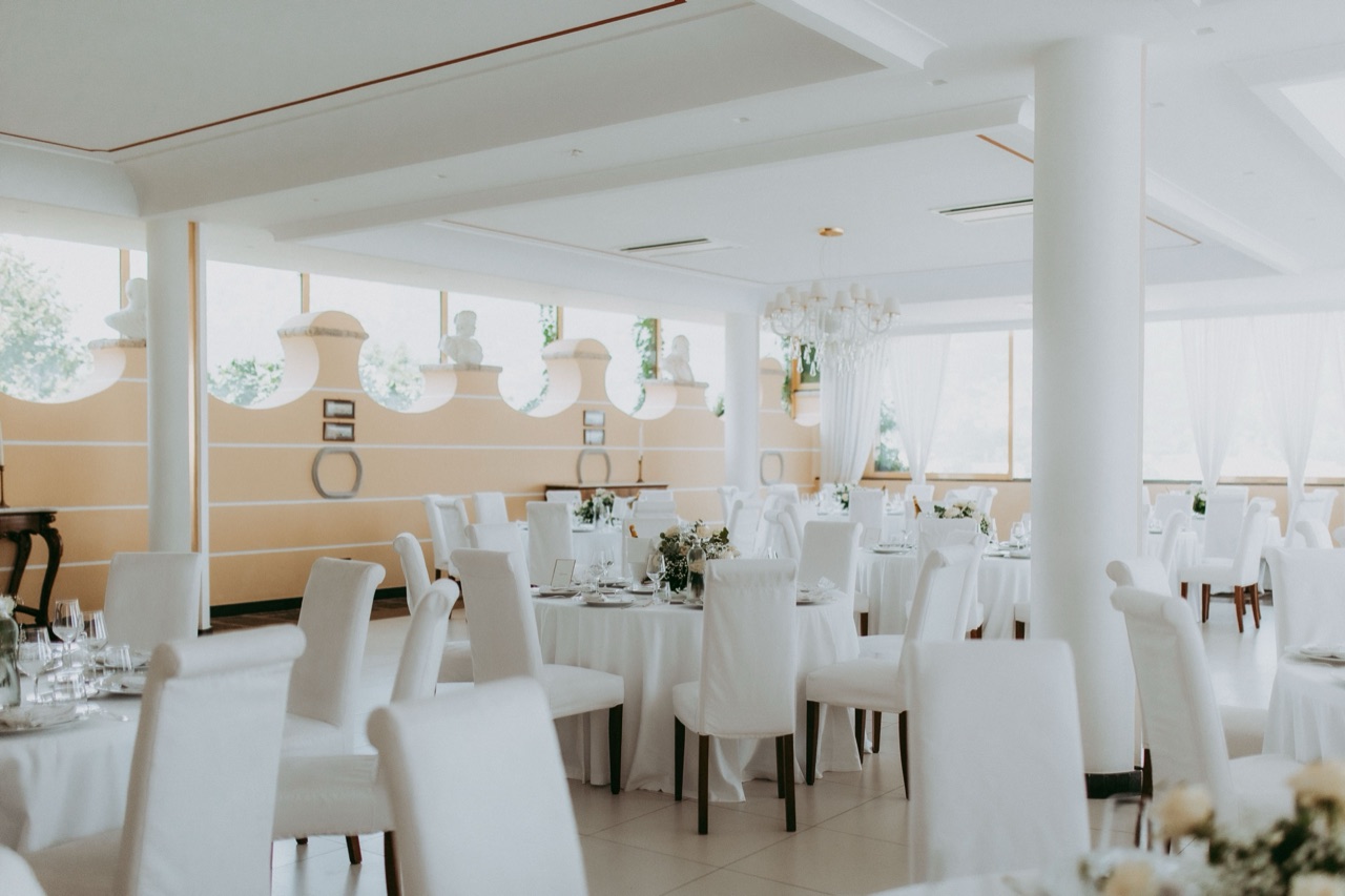 Sala Ricevimenti con tavoli rotondi, sedie vestite di bianco e lampadari di cristallo