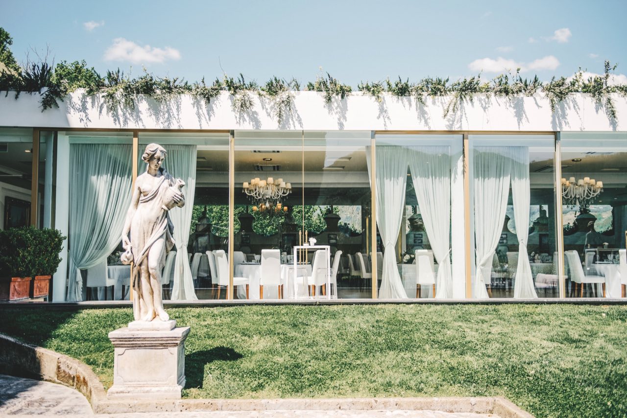 Sala Ricevimenti vista dal prato: parete vetrata con drappi chiari, festone di verde e statua in primo piano