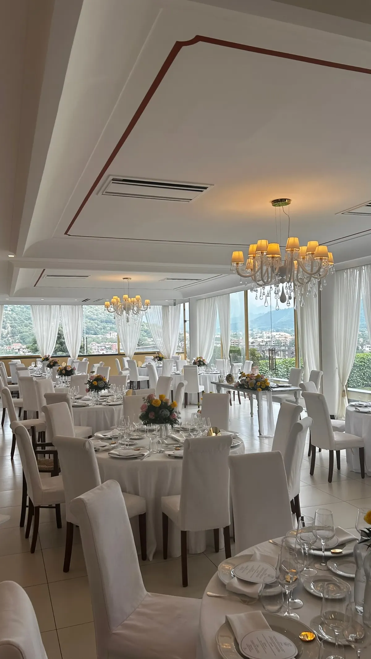 Sala Ricevimenti apparecchiata con tavoli rotondi, sedie vestite di bianco e lampadari di cristallo