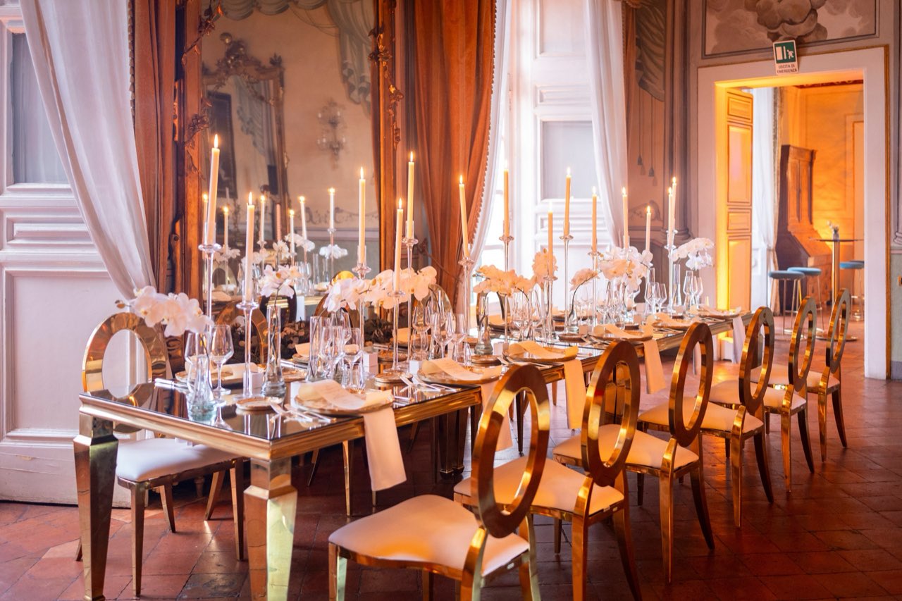 Tavolo imperiale con sedie dorate, candele alte e orchidee bianche in un salone storico affrescato